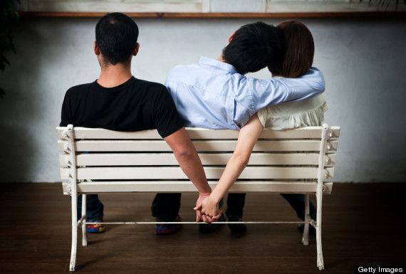 Two men and a woman sit on a bench. One man has his arm around the woman, while the woman holds hands with the other man behind his back.