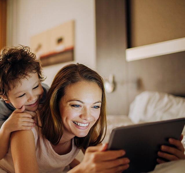 a mother lays on her stomach on a bed holding a tablet while her child looks over her shoulder